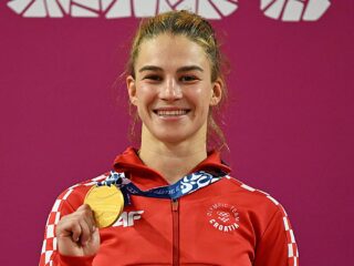 An athlete from Croatia smiles while holding a gold medal, wearing a red jacket with the Olympic Team Croatia logo.