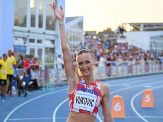 An athlete in a red and white Croatia uniform raises one arm in celebration on a blue track, with spectators visible in the background.